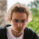 Young man with glasses and styled hair wearing a black hoodie, looking down thoughtfully outdoors.