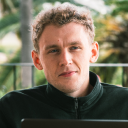 Portrait of a young man with curly hair wearing a black zip-up jacket, outdoors with blurred green foliage background.