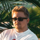 Man wearing black sunglasses and a white shirt, with palm leaves in the background.