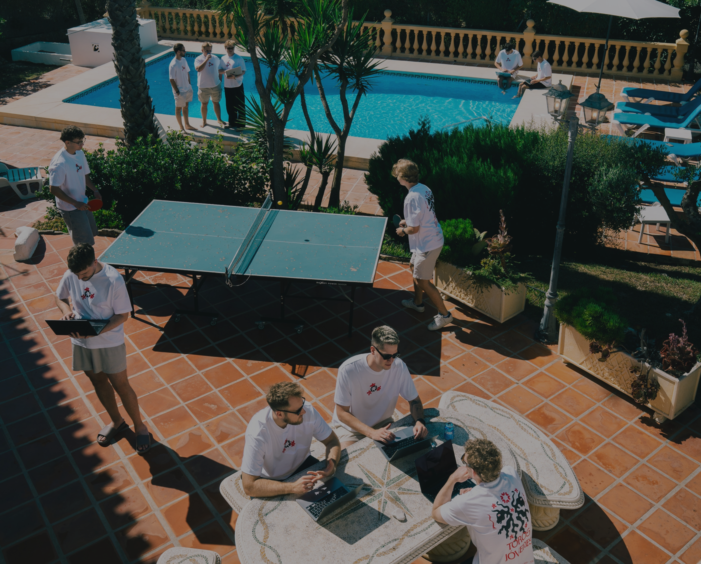 Group of young men in matching white shirts engaged in activities including playing table tennis, using laptops, and conversing near a poolside patio.