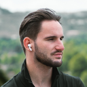 Side view of a young man with styled dark hair and a beard wearing white wireless earbuds outdoors.