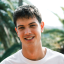 Smiling young man with short brown hair wearing a white shirt outdoors with blurred green foliage background.