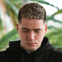 Young man with short curly hair looking down wearing a black hoodie with blurred outdoor greenery background.