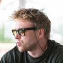 Man with disheveled hair wearing black sunglasses and earphones, looking contemplatively to the side.