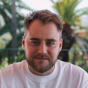 Man with short brown hair and beard wearing a white t-shirt, sitting outdoors with green plants in the background.