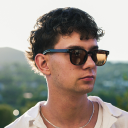 Young man with dark curly hair wearing black sunglasses and a white t-shirt outdoors.
